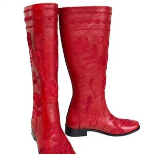 Puede incluir: Un par de botas altas de cuero rojo con intrincados bordados florales. Las botas presentan un dise&ntilde;o hasta la rodilla con punta redonda y tac&oacute;n bajo. El patr&oacute;n floral est&aacute; bordado en un tono m&aacute;s oscuro de rojo.