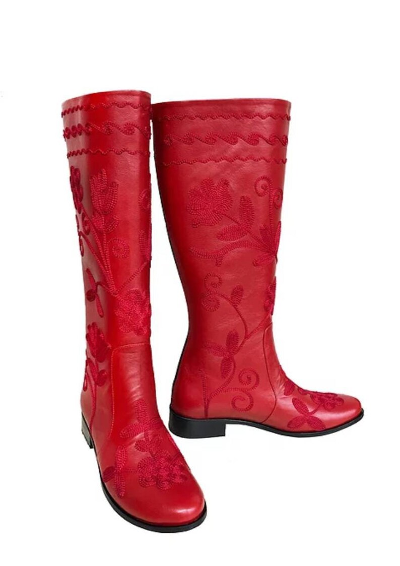 Puede incluir: Un par de botas altas de cuero rojo con intrincados bordados florales. Las botas presentan un dise&ntilde;o hasta la rodilla con punta redonda y tac&oacute;n bajo. El patr&oacute;n floral est&aacute; bordado en un tono m&aacute;s oscuro de rojo.
