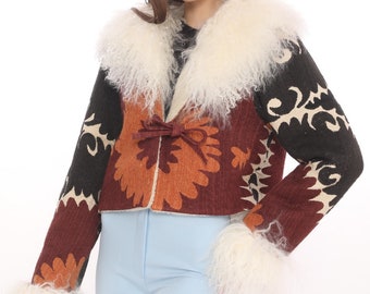 May include: A cropped jacket with a cream-colored fluffy collar and cuffs. The jacket features a floral design in shades of brown, orange, and black. The model is wearing light blue pants.