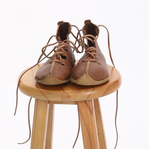 May include: A pair of brown leather boots with laces tied around a wooden stool. The boots have a pointed toe and are made of a textured leather.