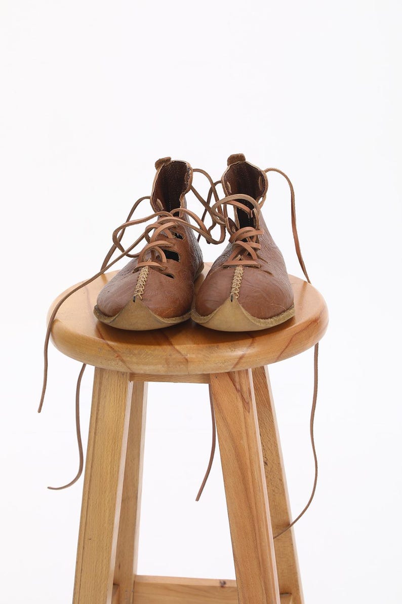 May include: A pair of brown leather boots with laces tied around a wooden stool. The boots have a pointed toe and are made of a textured leather.