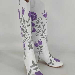 May include: White leather knee-high boots with a floral embroidery design. The flowers are purple and gray.