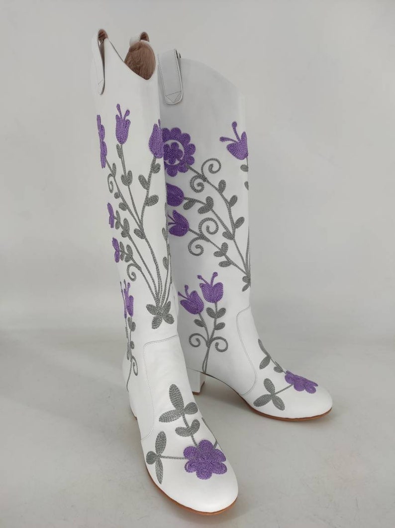 May include: White leather knee-high boots with a floral embroidery design. The flowers are purple and gray.