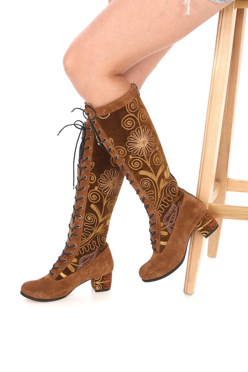 May include: Brown suede knee-high boots with a block heel and lace-up front. The boots feature intricate gold embroidery of floral designs.