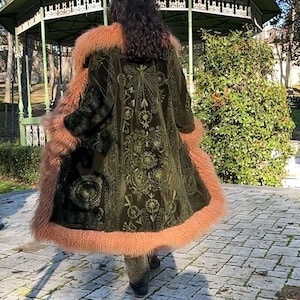 May include: A green velvet coat with a floral embroidered design and a pink faux fur trim. The coat is worn by a person standing in a park.
