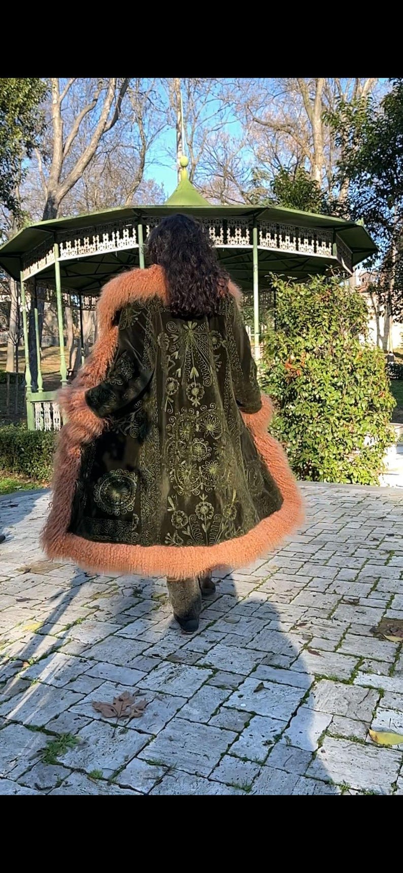 May include: A green velvet coat with a floral embroidered design and a pink faux fur trim. The coat is worn by a person standing in a park.