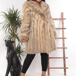 May include: A light brown fur coat with a wide collar and long sleeves. The coat is knee-length and has a natural, textured appearance. The model is wearing black leggings and black boots.