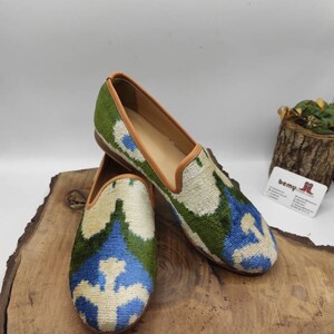 May include: A pair of green, blue, and white patterned velvet loafers with a brown leather trim. The loafers are sitting on a wooden surface.