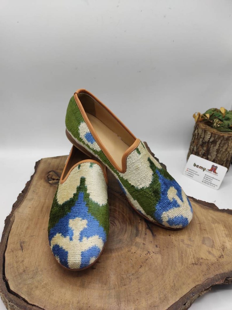 May include: A pair of green, blue, and white patterned velvet loafers with a brown leather trim. The loafers are sitting on a wooden surface.