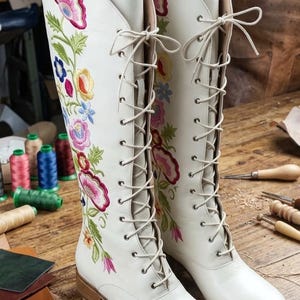 May include: A pair of white leather knee-high boots with floral embroidery in pink, yellow, blue, and green. The boots have a lace-up design and are set on a wooden surface, showcasing a handcrafted aesthetic.