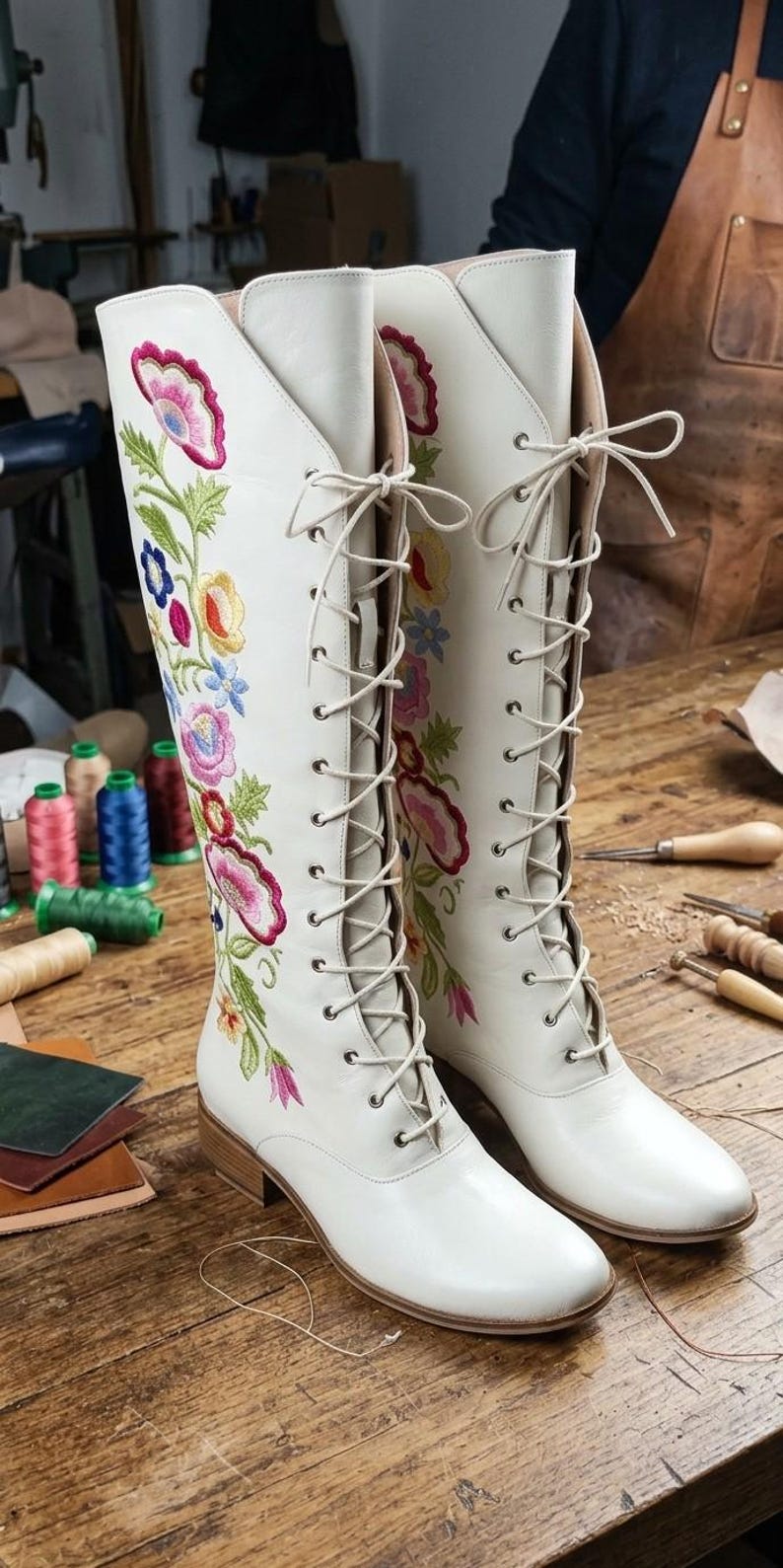 May include: A pair of white leather knee-high boots with floral embroidery in pink, yellow, blue, and green. The boots have a lace-up design and are set on a wooden surface, showcasing a handcrafted aesthetic.