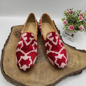 May include: A pair of red and white patterned velvet loafers with a brown leather trim. The loafers are on a wooden surface.