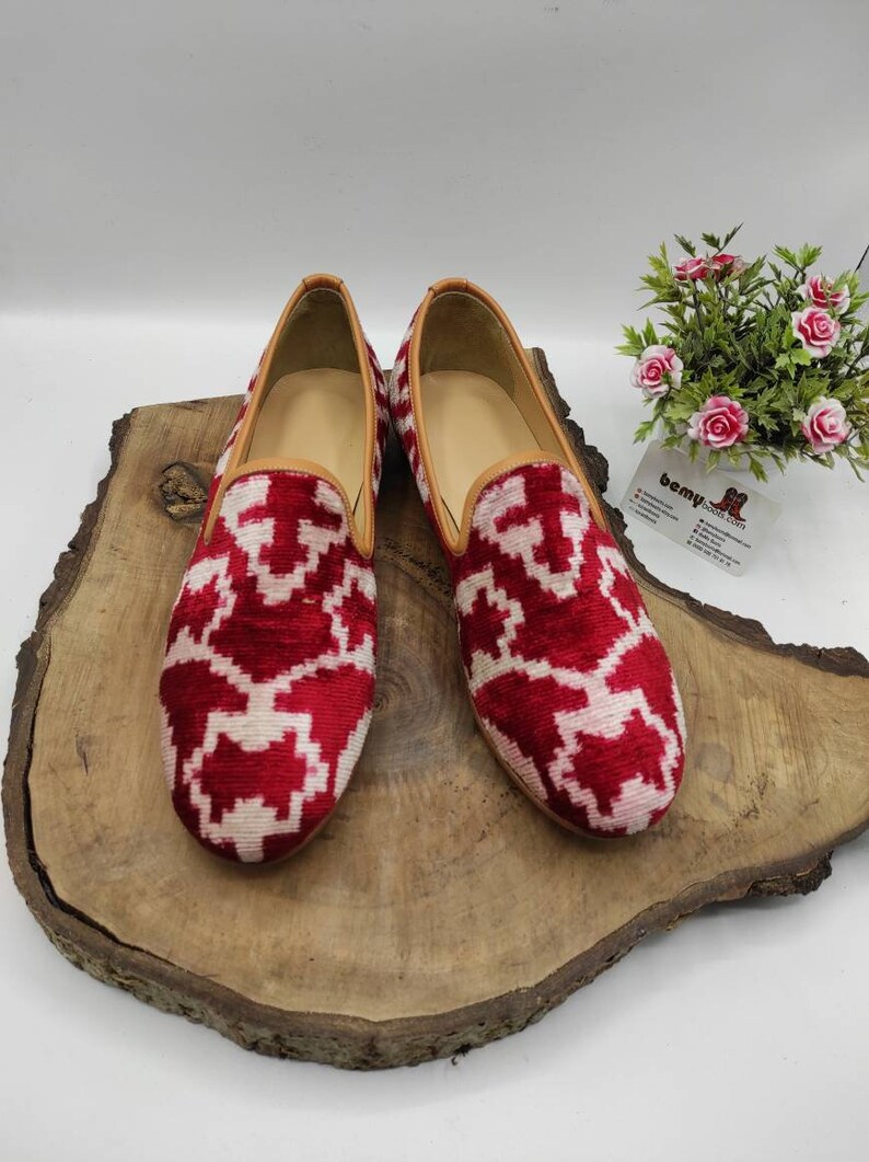 May include: A pair of red and white patterned velvet loafers with a brown leather trim. The loafers are on a wooden surface.