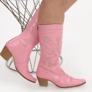 May include: A pair of pink cowboy boots with floral embroidery. The boots have a pointed toe and a low heel.
