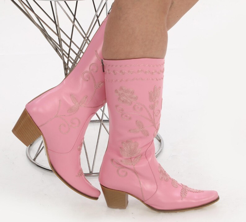 May include: A pair of pink cowboy boots with floral embroidery. The boots have a pointed toe and a low heel.