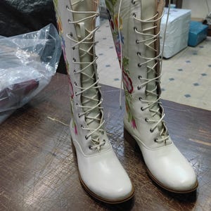 May include: A pair of white knee-high boots with floral embroidery. The boots have a lace-up design and a small heel. The floral embroidery features pink, blue, and yellow flowers. The boots are made of a smooth, shiny material.