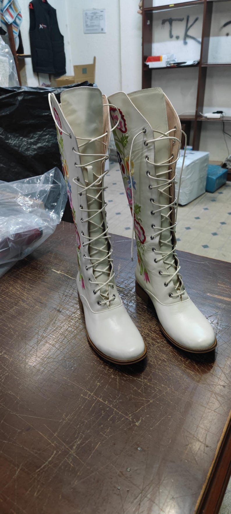 May include: A pair of white knee-high boots with floral embroidery. The boots have a lace-up design and a small heel. The floral embroidery features pink, blue, and yellow flowers. The boots are made of a smooth, shiny material.