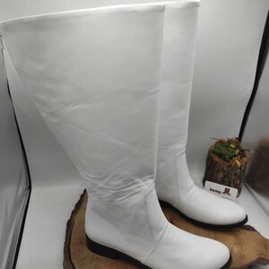 Puede incluir: Un par de botas blancas de cuero de ca&ntilde;a alta con tac&oacute;n bajo. Las botas est&aacute;n sobre una superficie de madera.