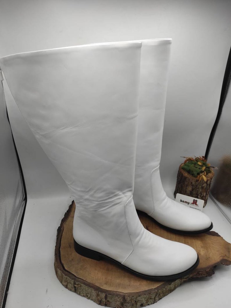Puede incluir: Un par de botas blancas de cuero de ca&ntilde;a alta con tac&oacute;n bajo. Las botas est&aacute;n sobre una superficie de madera.