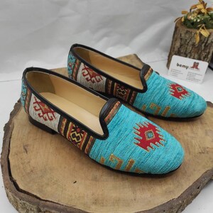 May include: A pair of turquoise slip-on shoes with a woven design. The shoes feature a black leather trim and a colorful geometric pattern in red, yellow, and brown. The shoes are displayed on a wooden surface.
