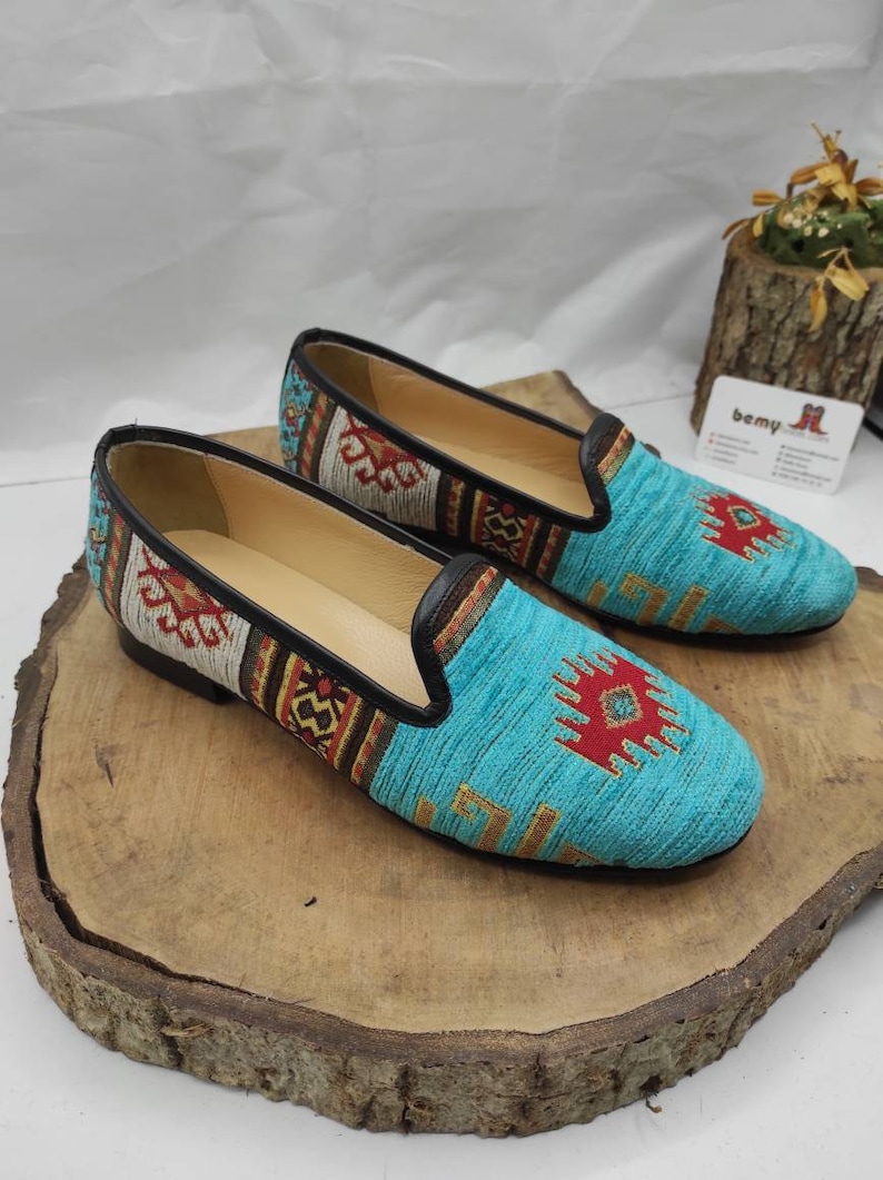 May include: A pair of turquoise slip-on shoes with a woven design. The shoes feature a black leather trim and a colorful geometric pattern in red, yellow, and brown. The shoes are displayed on a wooden surface.