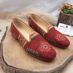 May include: A pair of red slip-on shoes with a woven textile design. The shoes have a tan leather trim and a decorative pattern in shades of beige, gray, and red. The shoes are displayed on a wooden surface.