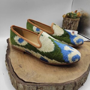 May include: A pair of green, blue, and white patterned velvet loafers with brown leather trim. The loafers are sitting on a wooden surface.