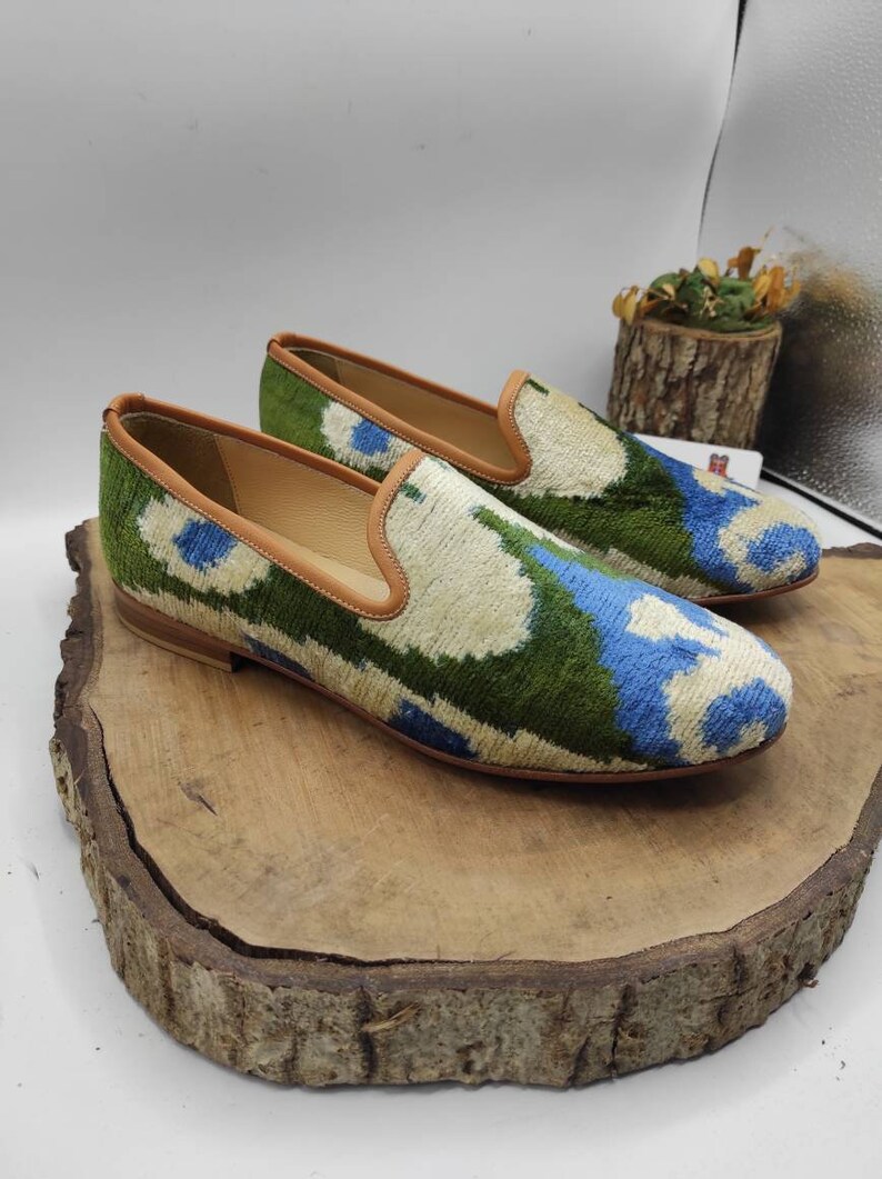May include: A pair of green, blue, and white patterned velvet loafers with brown leather trim. The loafers are sitting on a wooden surface.