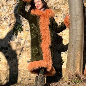 May include: A woman wearing a long, green velvet coat with a pink fur trim. The coat has an embroidered floral pattern. She is standing in front of a stone wall and a tree.