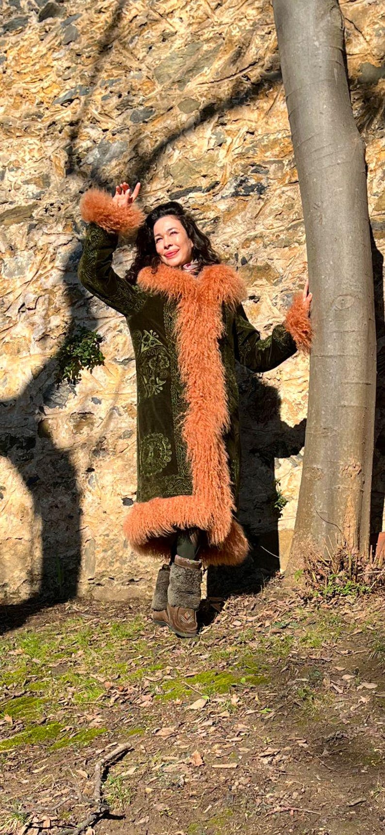 May include: A woman wearing a long, green velvet coat with a pink fur trim. The coat has an embroidered floral pattern. She is standing in front of a stone wall and a tree.