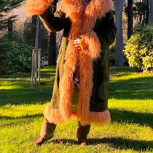 May include: A woman wearing a long, green velvet coat with a fluffy, peach-colored fur collar and trim. The coat has a paisley pattern and is worn over a brown turtleneck sweater. The woman is standing in a grassy park with trees in the background.