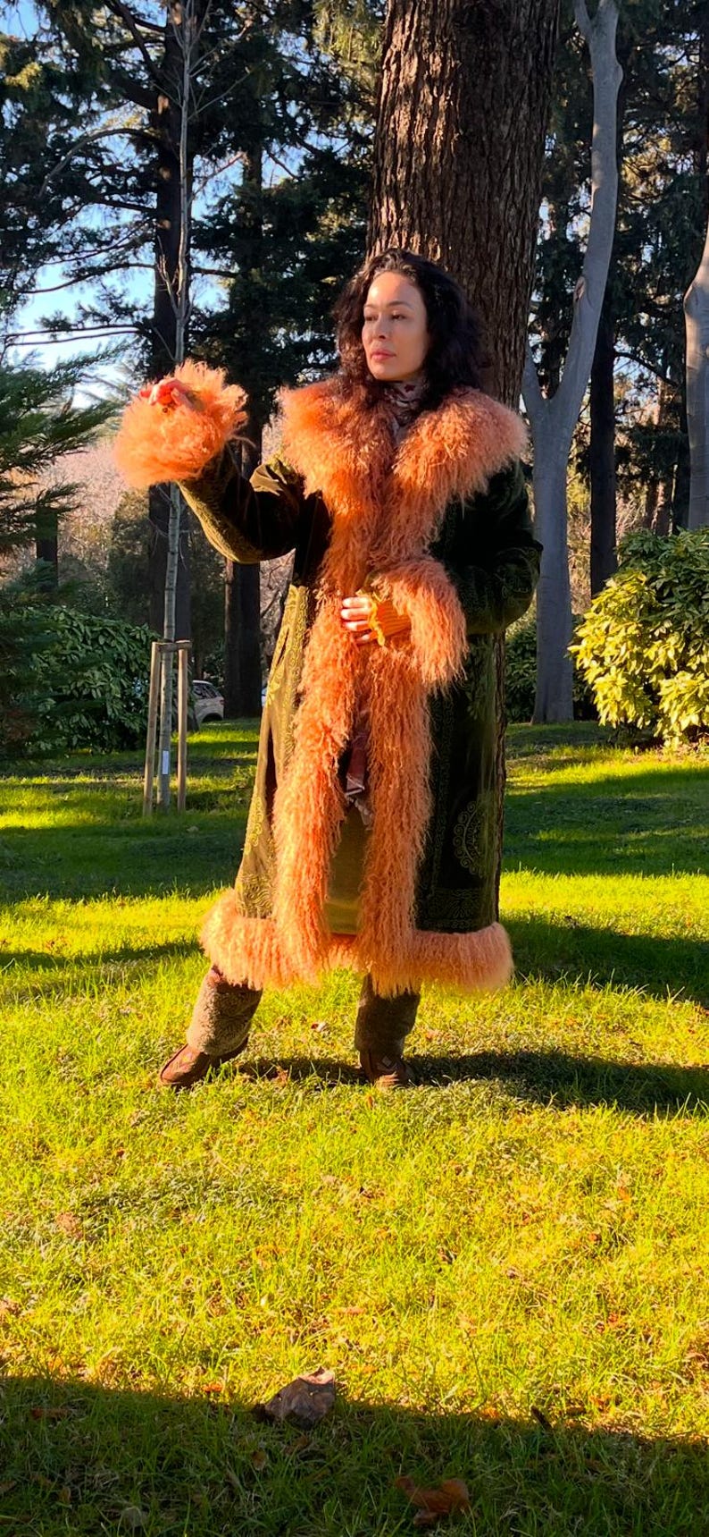 May include: A woman wearing a long, green velvet coat with a fluffy, peach-colored fur collar and trim. The coat has a paisley pattern and is worn over a brown turtleneck sweater. The woman is standing in a grassy park with trees in the background.