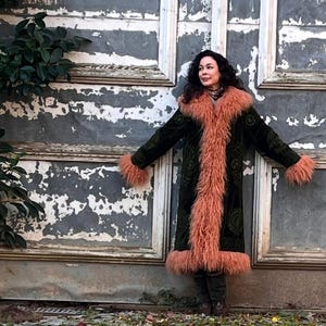 May include: A woman wearing a long, dark green velvet coat with a pink faux fur trim. The coat has a high collar and is lined with a pink faux fur. The woman is standing in front of a weathered wooden wall.
