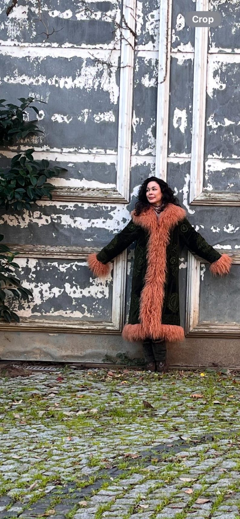 May include: A woman wearing a long, dark green velvet coat with a pink faux fur trim. The coat has a high collar and is lined with a pink faux fur. The woman is standing in front of a weathered wooden wall.