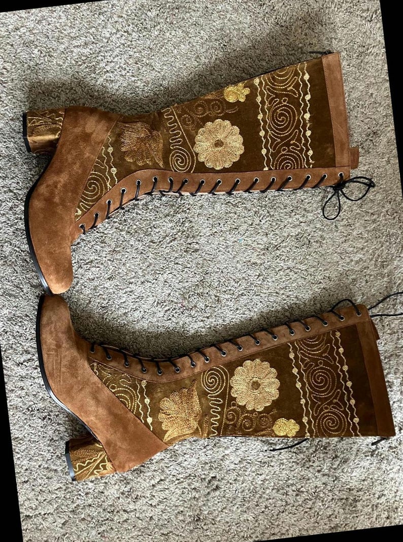 May include: A pair of brown suede knee-high boots with gold embroidery and lace-up closures. The boots have a block heel.