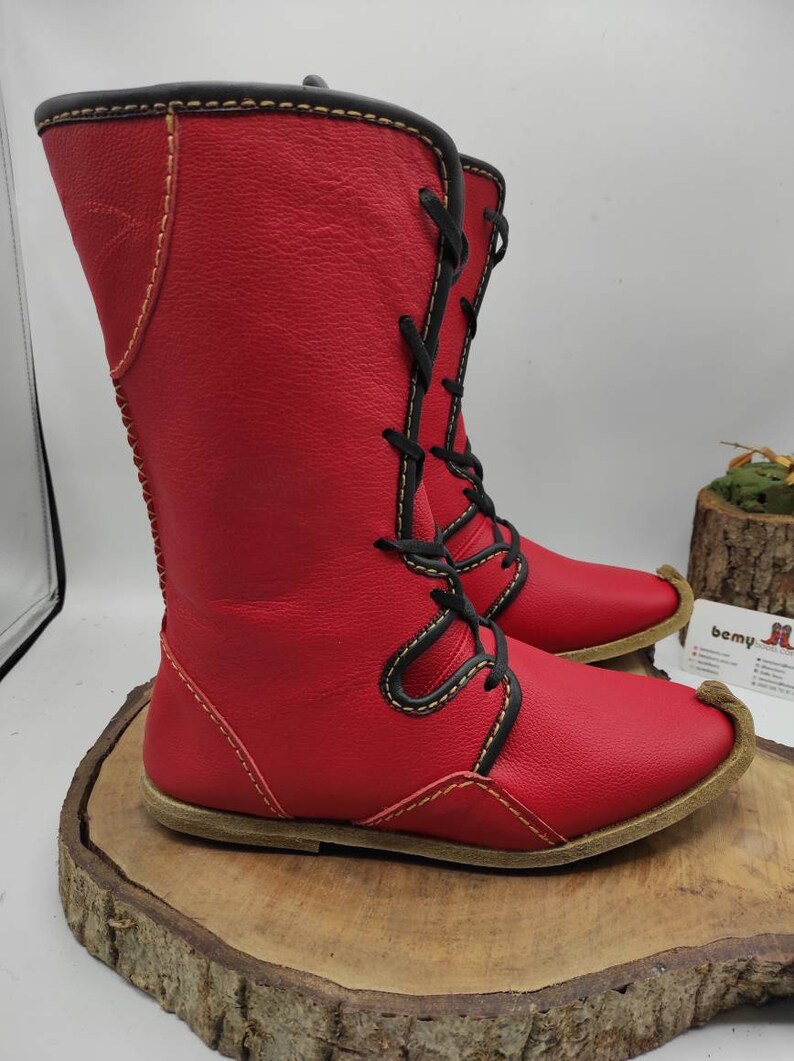 May include: Red leather boots with black laces and yellow stitching. The boots have a pointed toe and a flat sole.
