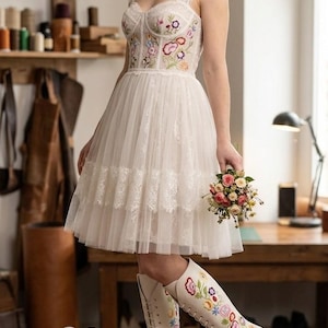 May include: A white wedding dress with a floral embroidered bodice and a tulle skirt. The dress is paired with matching white knee-high boots, also adorned with floral embroidery. A small bouquet of flowers is held in the hand.