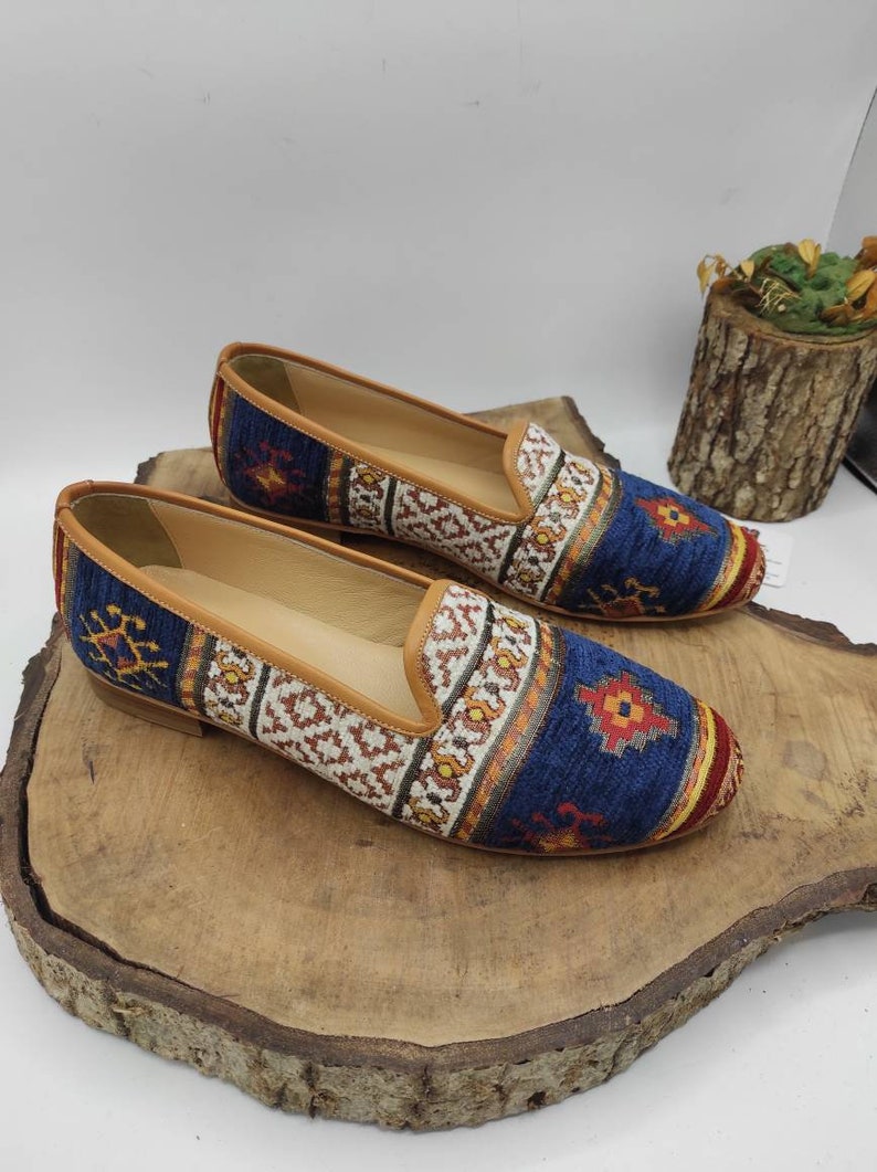 May include: A pair of flat, slip-on shoes with a patterned textile upper. The shoes feature a blue background with geometric designs in red, orange, and white. The trim and inner sole are tan, and the soles are brown.