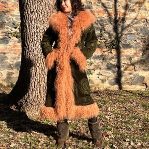 May include: A long, green velvet coat with a fluffy, peach-colored faux fur collar and trim. The coat is open and the fur is visible. The person wearing the coat is standing in front of a brick wall and a tree.