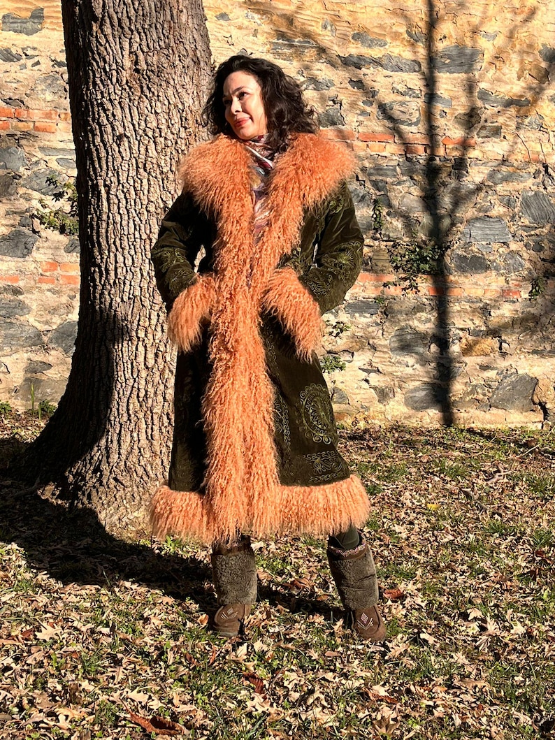 May include: A long, green velvet coat with a fluffy, peach-colored faux fur collar and trim. The coat is open and the fur is visible. The person wearing the coat is standing in front of a brick wall and a tree.
