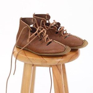 May include: A pair of brown leather sandals with laces. The sandals are sitting on a wooden stool.