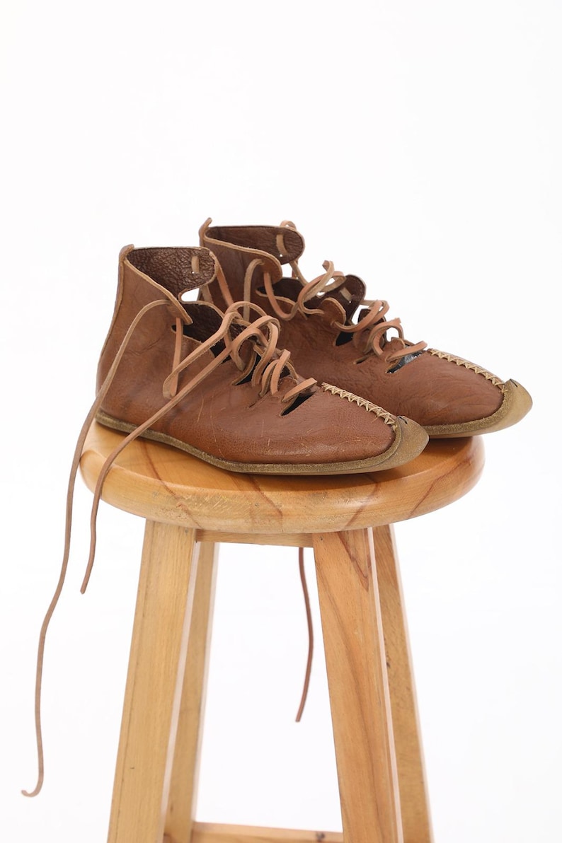 May include: A pair of brown leather sandals with laces. The sandals are sitting on a wooden stool.