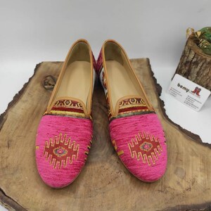 May include: A pair of pink slip-on shoes with a patterned design on the toe and upper. The shoes have a tan leather trim and a light-colored interior. The shoes are displayed on a wooden surface.