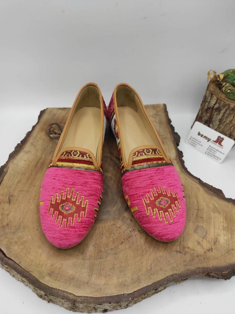 May include: A pair of pink slip-on shoes with a patterned design on the toe and upper. The shoes have a tan leather trim and a light-colored interior. The shoes are displayed on a wooden surface.