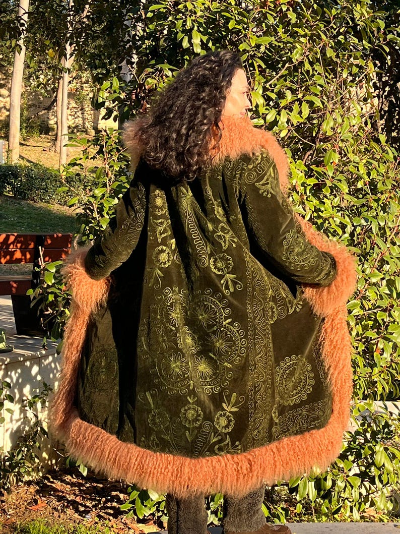 May include: A long, green velvet coat with a floral pattern and a fluffy, peach-colored fur trim. The coat has a hood and is lined with fur.