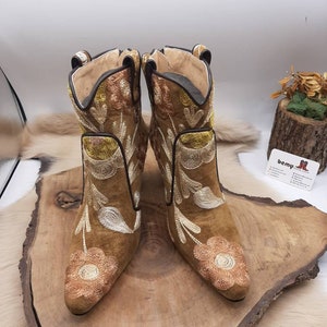 May include: A pair of brown suede ankle boots with floral embroidery in shades of gold, white, and brown. The boots have a pointed toe and a low heel.