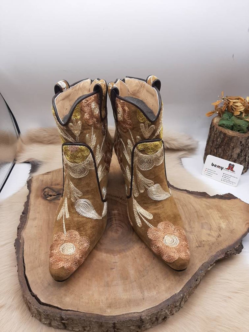 May include: A pair of brown suede ankle boots with floral embroidery in shades of gold, white, and brown. The boots have a pointed toe and a low heel.