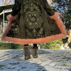 May include: A green velvet coat with embroidered floral designs and a pink faux fur trim. The coat is worn by a person standing on a stone path.