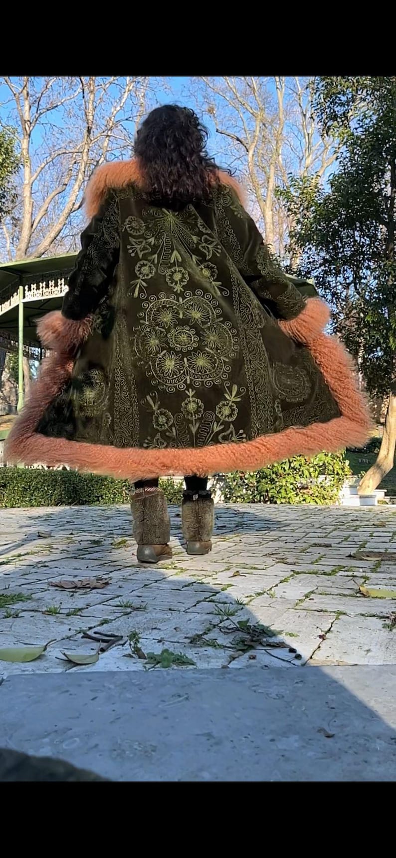 May include: A green velvet coat with embroidered floral designs and a pink faux fur trim. The coat is worn by a person standing on a stone path.