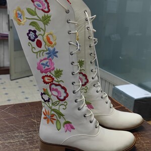 May include: White knee-high boots with colorful floral embroidery. The boots feature a lace-up design and a low heel. The floral design includes pink, blue, yellow, and orange flowers with green leaves. The boots are displayed on a wooden surface.